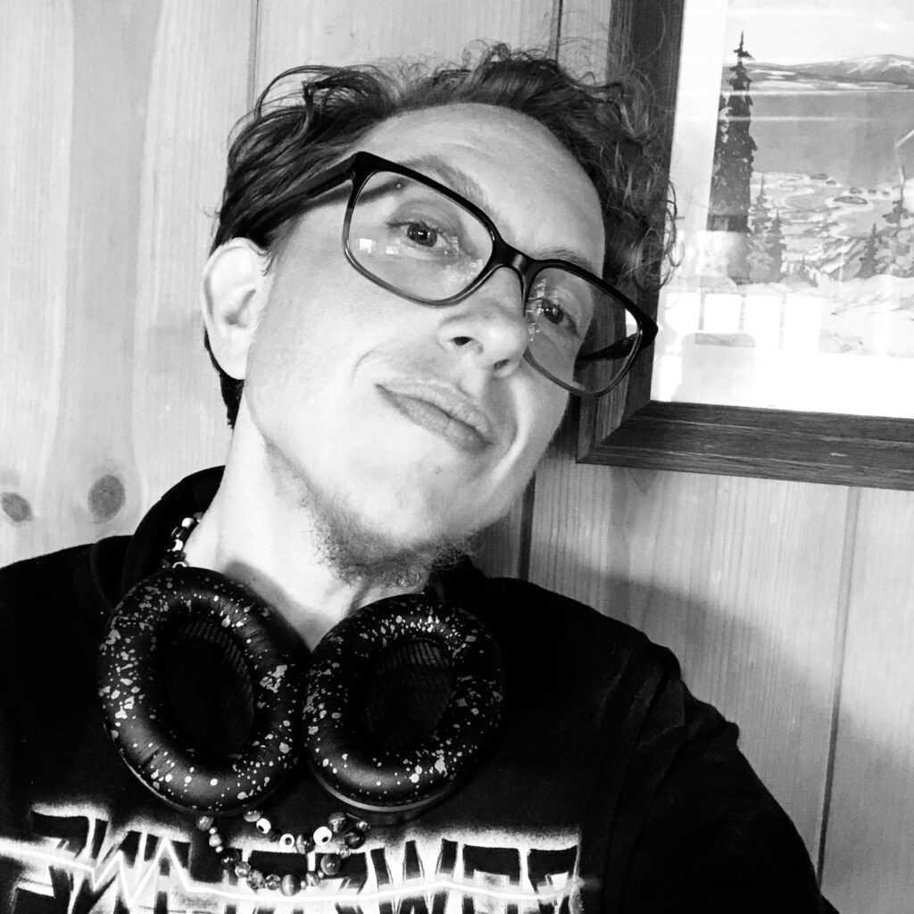 A black and white image of M. Lopes da Silva leaning back against a wooden wall and a watercolor of a lighthouse. He is a white trans masc individual with blonde curly hair, large black glasses, a black shirt, and black-and-white speckled headphones around his neck.